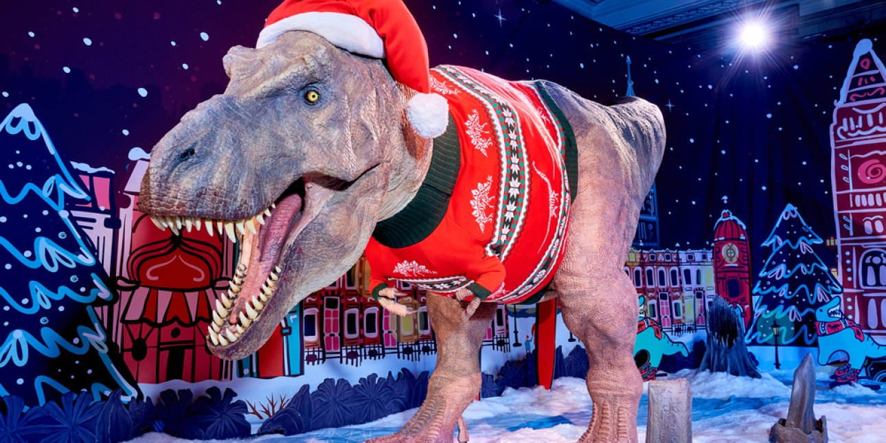A large animatronic T-rex wearing a festive jumper and Santa hat. The background is an illustration of snow covered buildings